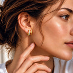 A close-up lifestyle shot of a woman wearing a small gold huggie hoop earring with a triple-stripe design of white, yellow, and green diamonds.
