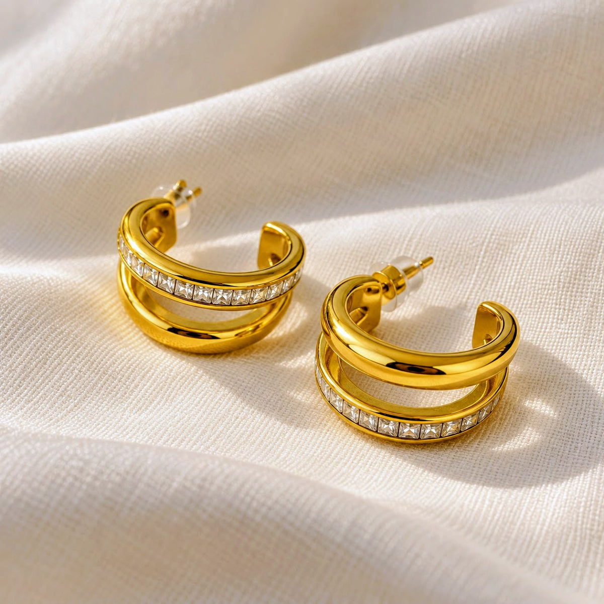 A pair of gold hoop earrings with a double-band design and a row of clear stones on the front, displayed on a draped cream-colored fabric background.