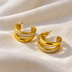 A pair of gold hoop earrings with a double-band design and a row of clear stones on the front, displayed on a draped cream-colored fabric background.