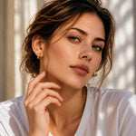A close-up portrait of a woman wearing small gold hoop earrings, with warm sunlight and soft shadows across her face and white top.