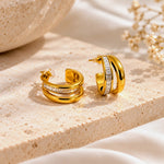 A pair of gold hoop earrings with a double-band design and a row of clear stones on the front, displayed on a draped cream-colored fabric background.