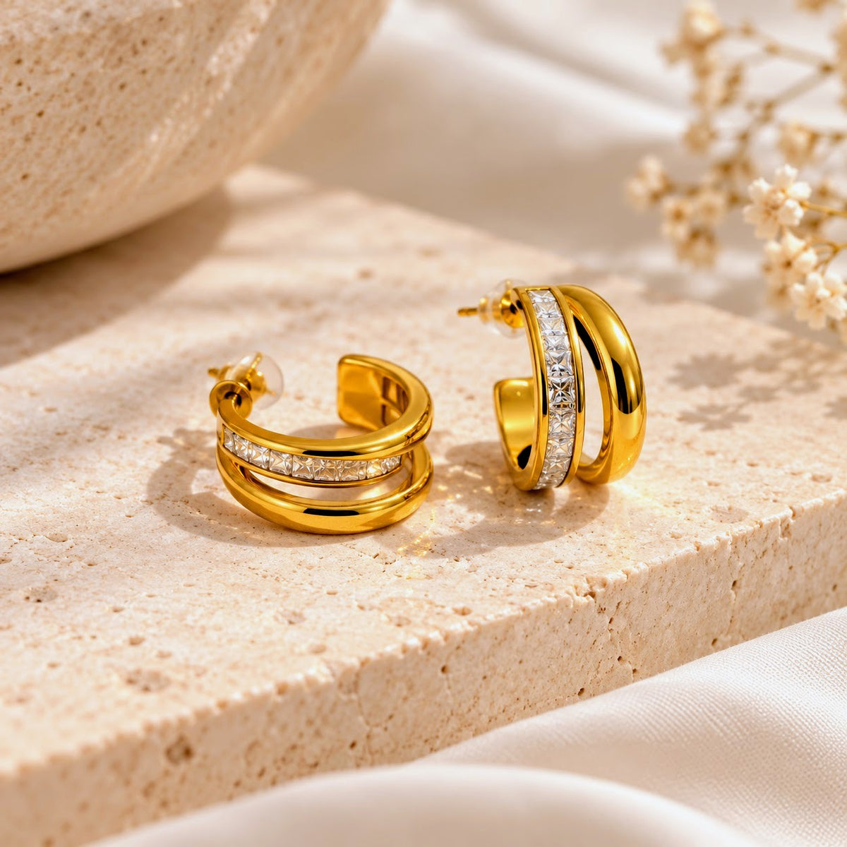 A pair of gold hoop earrings with a double-band design and a row of clear stones on the front, displayed on a draped cream-colored fabric background.