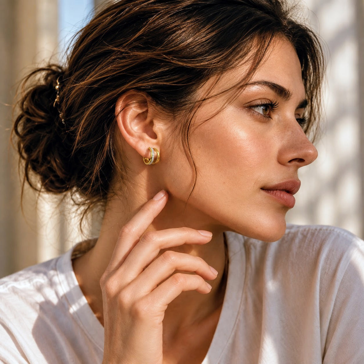 A close-up lifestyle shot of a woman with her hair in a bun, wearing a small gold crescent moon stud earring and a white top, set against a softly lit, sun-dappled beige background.