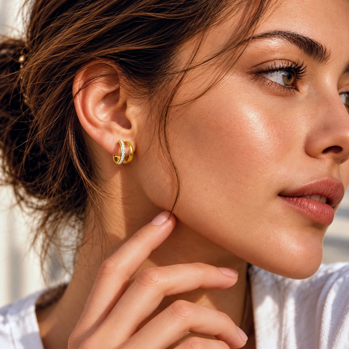 A close-up lifestyle shot of a woman wearing a small gold hoop earring with clear gemstones on her earlobe.
