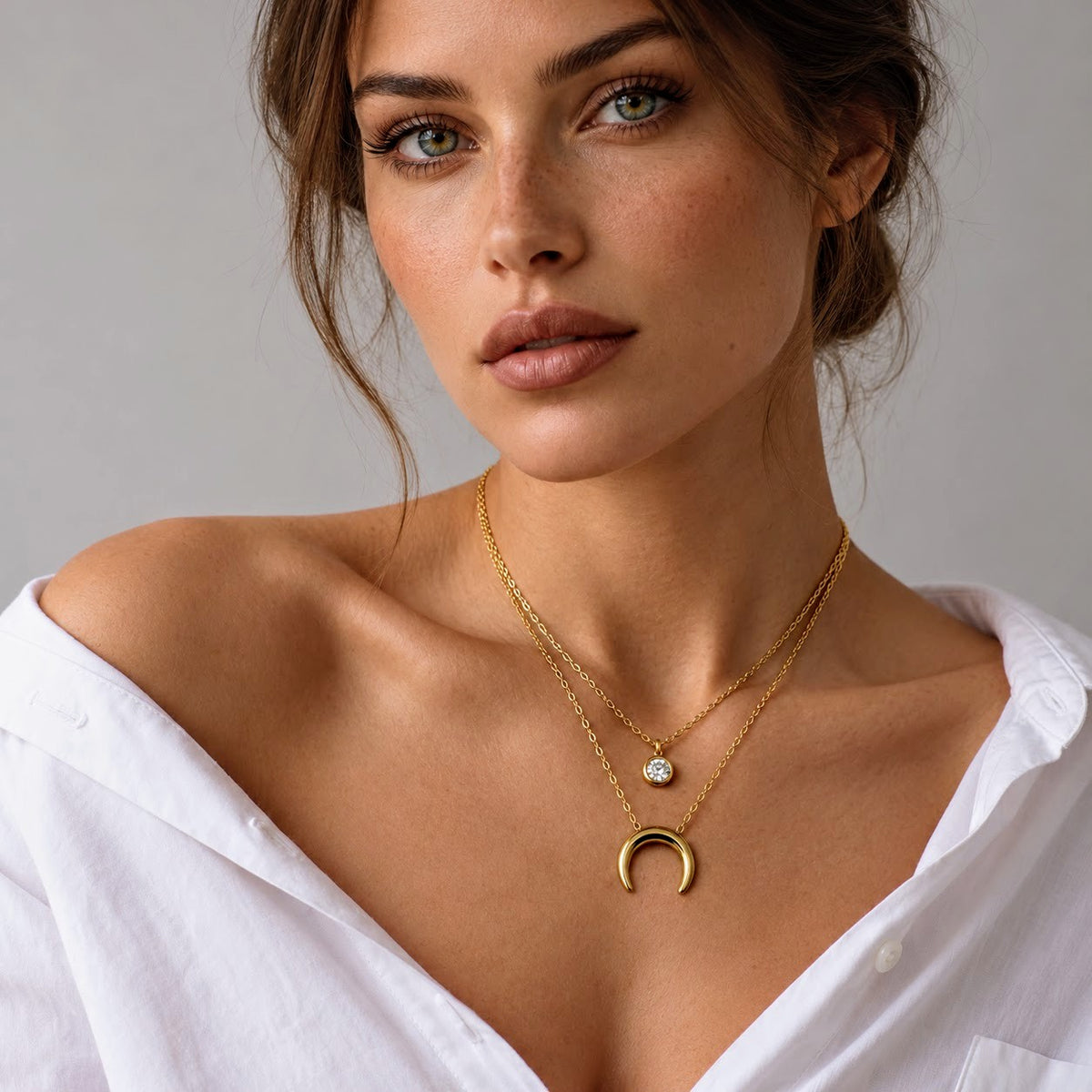 A close-up of a woman wearing a double-layered gold necklace with a crescent moon pendant and a small round stone charm, paired with an off-the-shoulder white shirt against a neutral gray background.