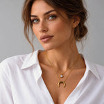 A close-up of a woman wearing a double-layered gold necklace with a crescent moon pendant and a small round stone charm, paired with a white collared shirt against a neutral gray background.