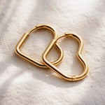 Two gold-toned metal heart-shaped hoop earrings resting on a textured off-white fabric background.