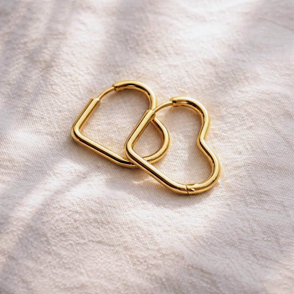 Two gold heart-shaped hoop earrings overlapping on a textured off-white fabric background with soft shadows.