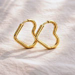 A pair of gold-plated geometric hoop earrings with a wavy, open heart-like shape, resting on a textured off-white fabric background.