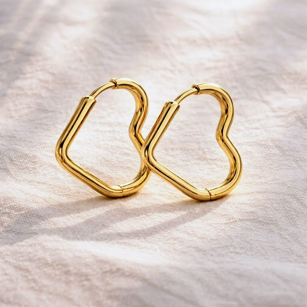 A pair of gold-plated geometric hoop earrings with a wavy, open heart-like shape, resting on a textured off-white fabric background.
