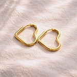 A pair of gold heart-shaped hoop earrings resting on a textured, light beige fabric background.