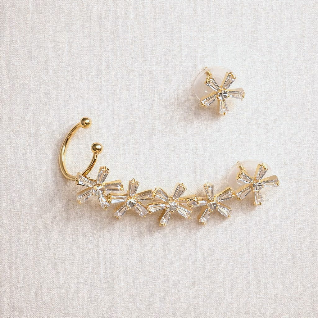 Gold-toned curved ear cuff jewelry with five clear faceted crystal flower motifs, shown on a light beige textured fabric background alongside a matching clear crystal stud earring.