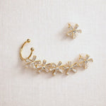 Gold-toned curved ear cuff jewelry with five clear faceted crystal flower motifs, shown on a light beige textured fabric background alongside a matching clear crystal stud earring.