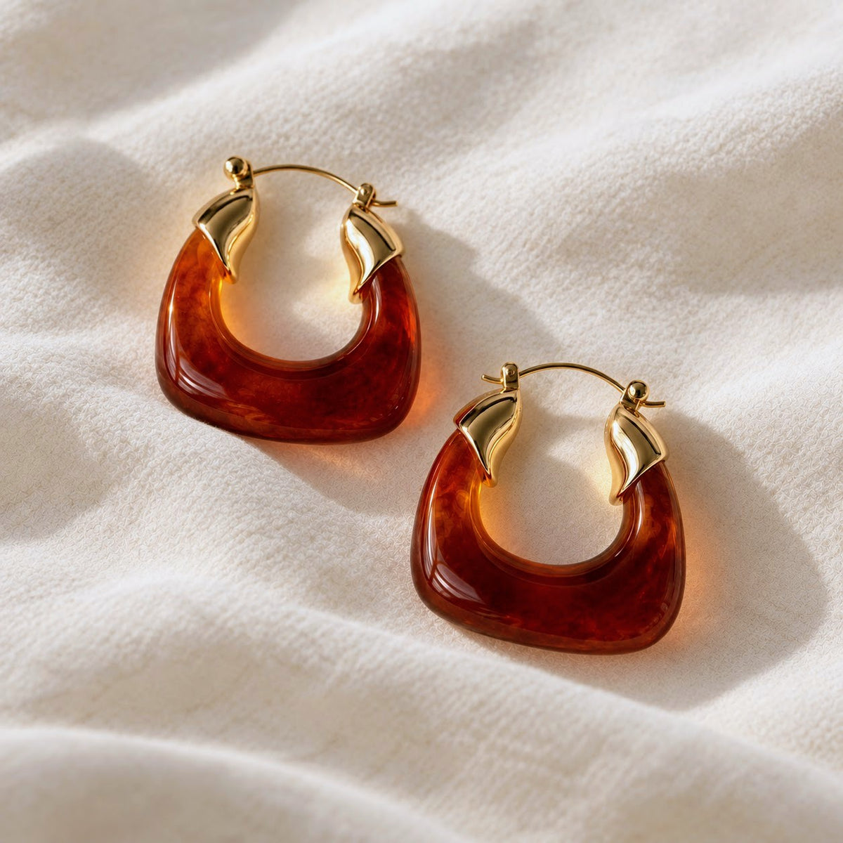 A pair of chunky square-shaped hoop earrings with a translucent amber-brown resin body and polished gold-tone metal posts, displayed on a textured white fabric background.