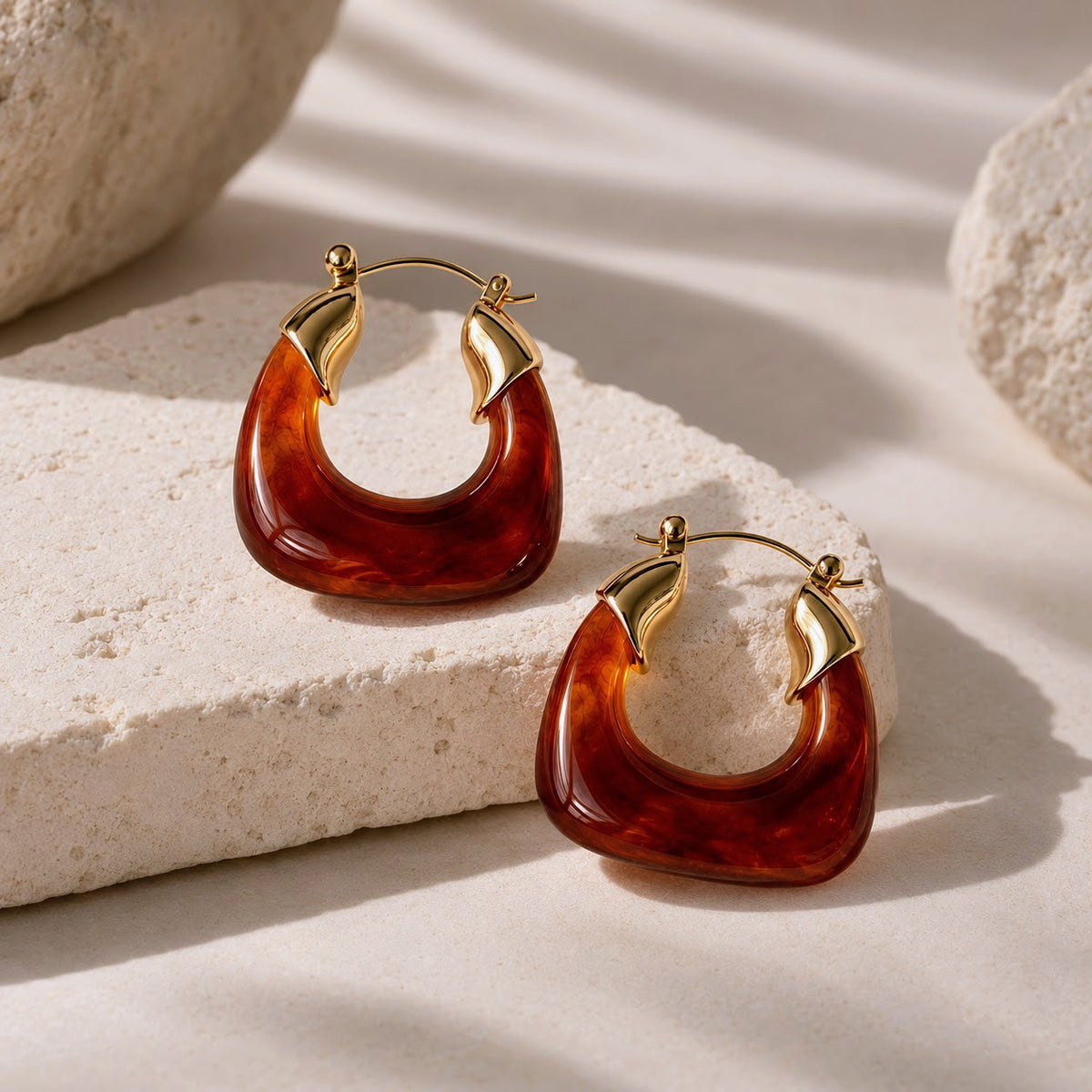 A pair of chunky crescent-shaped hoop earrings made of glossy amber-brown translucent resin with gold-tone metal hoops and leaf-like accents, displayed on a textured light stone surface with soft shadows.