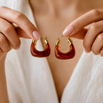 A close-up lifestyle shot of a person holding a pair of chunky, crescent-shaped hoop earrings. The earrings feature a glossy, translucent reddish-brown resin body with polished gold metal leaf accents at the top, held against a soft white top.