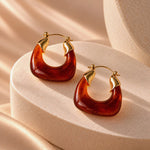 A pair of gold hoop earrings with curved amber-brown resin inserts, displayed on a beige textured pedestal with soft warm lighting.