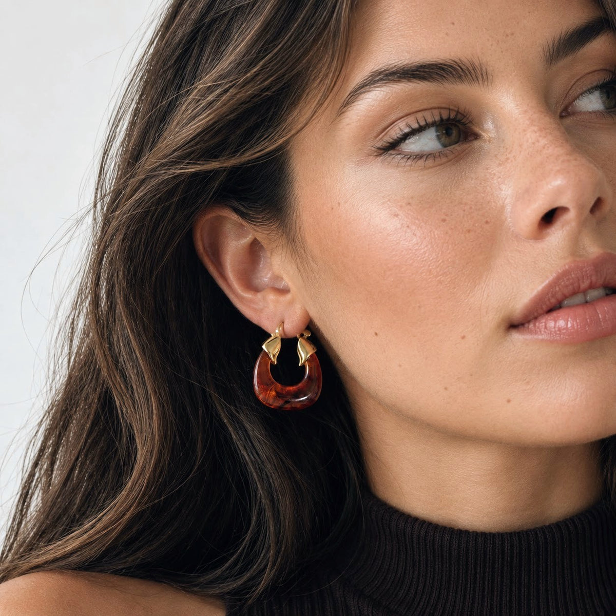 A close-up of a woman wearing a pair of gold crescent-shaped hoop earrings with amber-brown translucent drop charms, set against a clean light gray background.