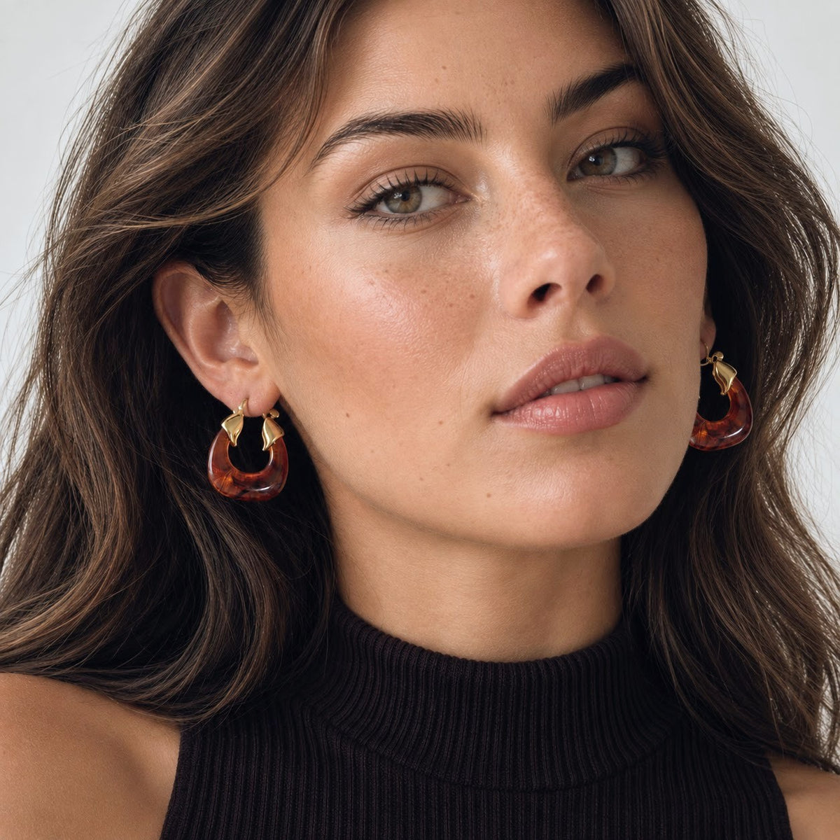A close-up of a woman wearing large, chunky gold hoop earrings with translucent amber-brown inner sections, paired with a black ribbed turtleneck top against a light gray background.