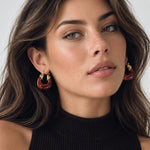 A close-up of a woman wearing large, chunky gold hoop earrings with translucent amber-brown inner sections, paired with a black ribbed turtleneck top against a light gray background.