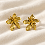 A pair of textured gold flower stud earrings with a lily design, displayed on a soft, draped cream-colored fabric background.