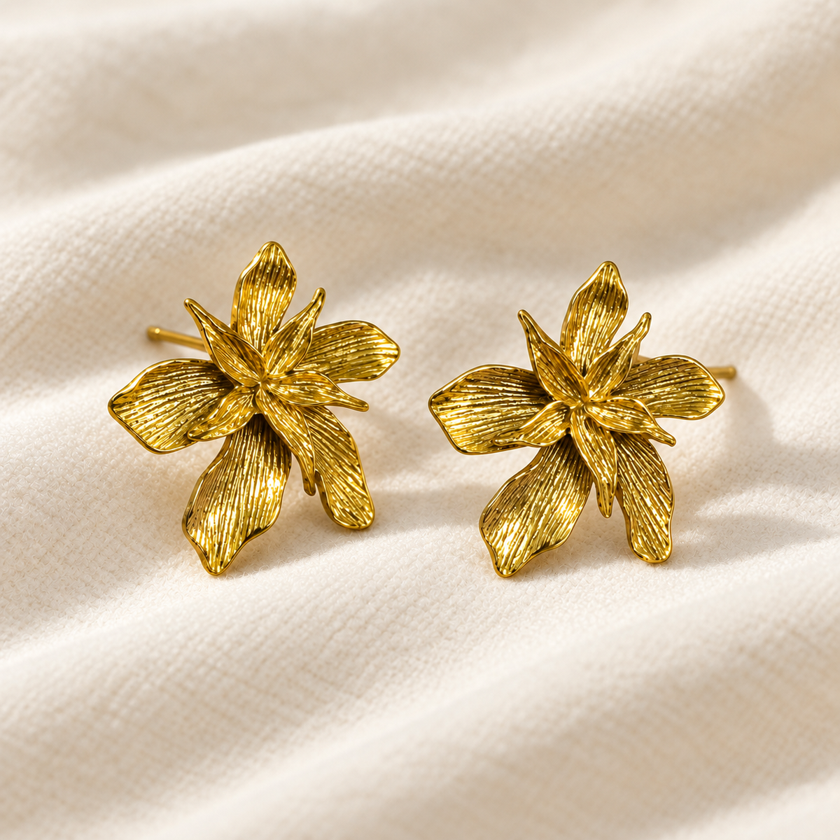 A pair of textured gold flower stud earrings with a lily design, displayed on a soft, draped cream-colored fabric background.