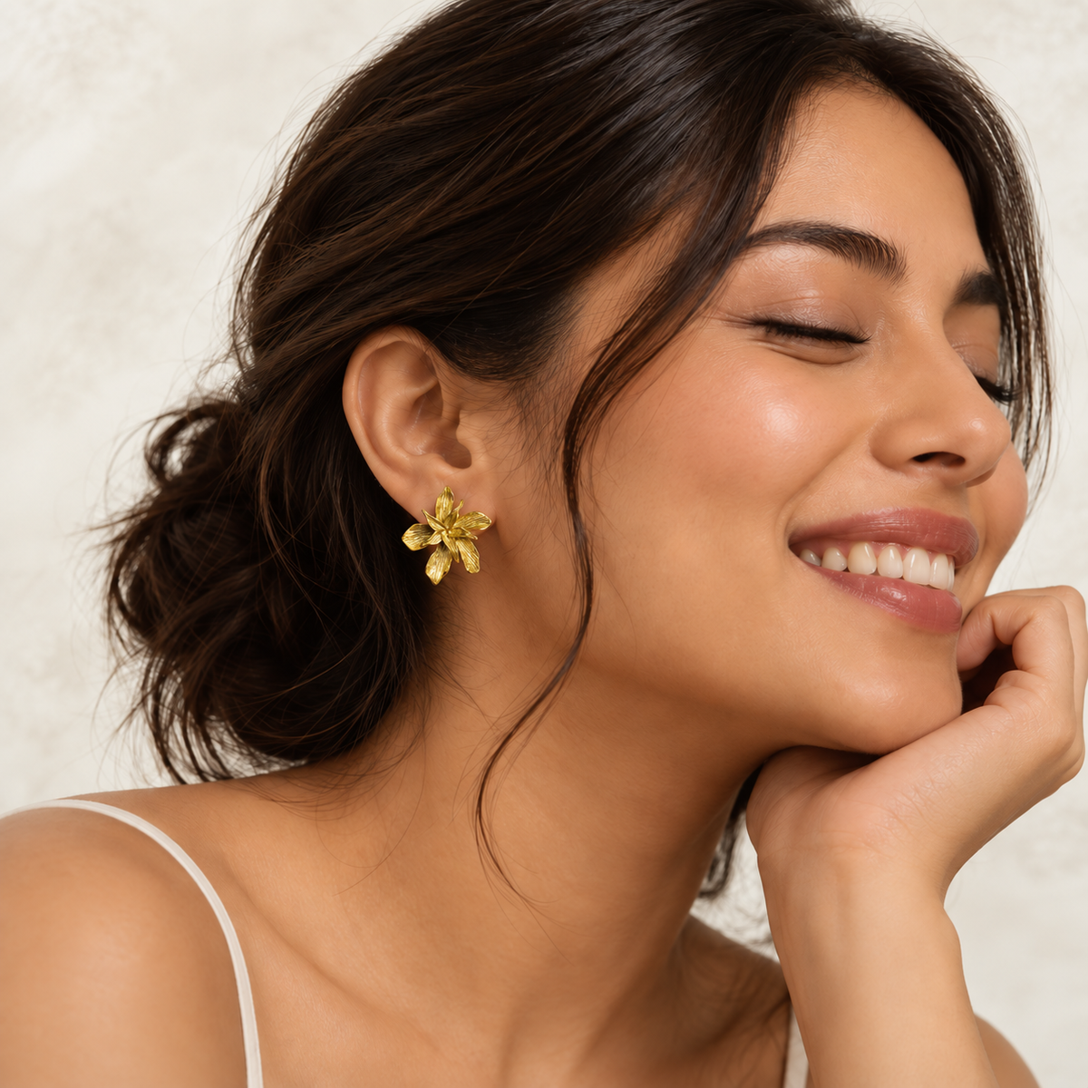 A close-up lifestyle shot of a woman with dark hair in an updo, wearing a textured gold flower-shaped stud earring. She is smiling with her eyes closed, resting her chin on her hand against a soft, light beige textured background.