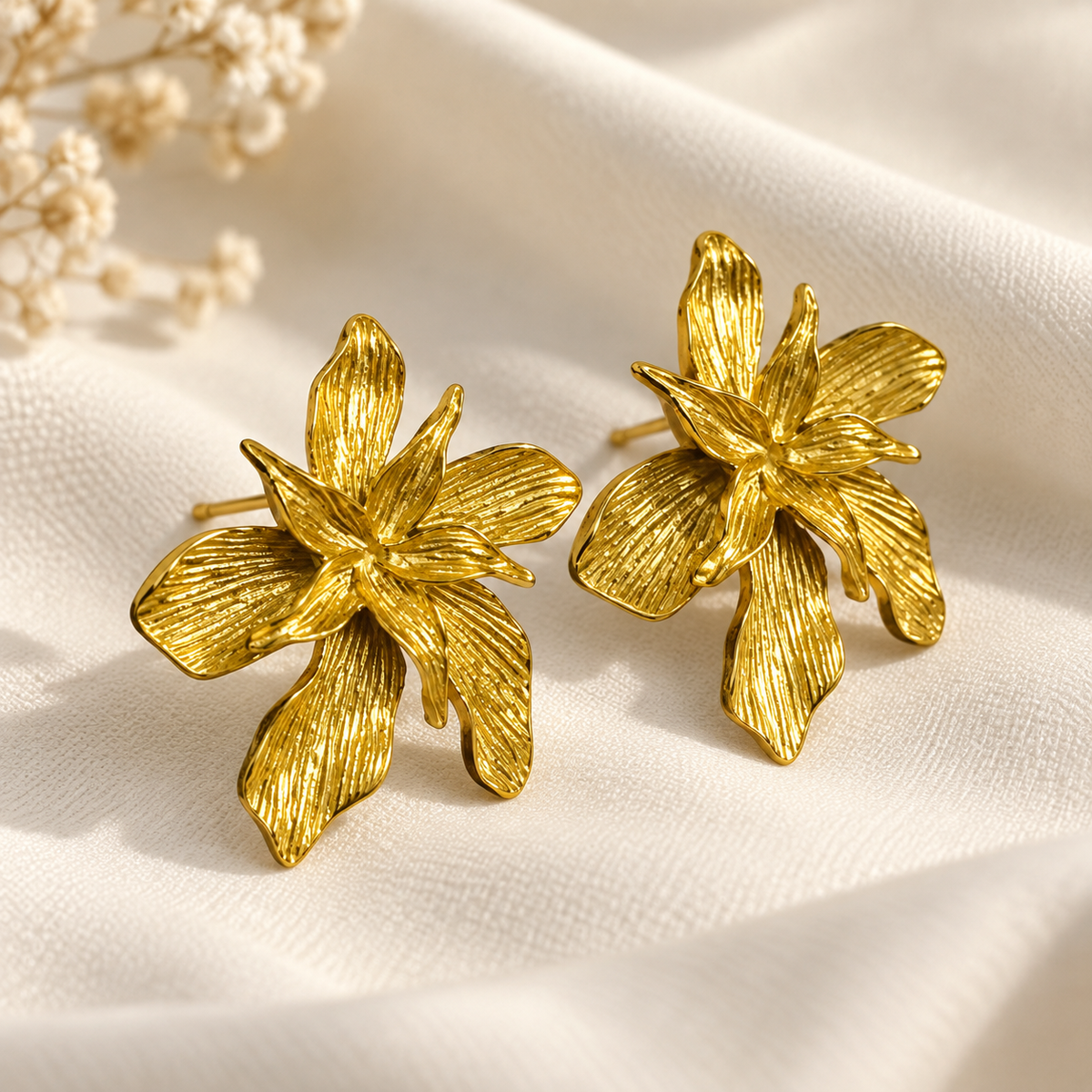 A pair of textured gold flower stud earrings displayed on a soft, draped cream fabric with small dried white flowers in the corner.