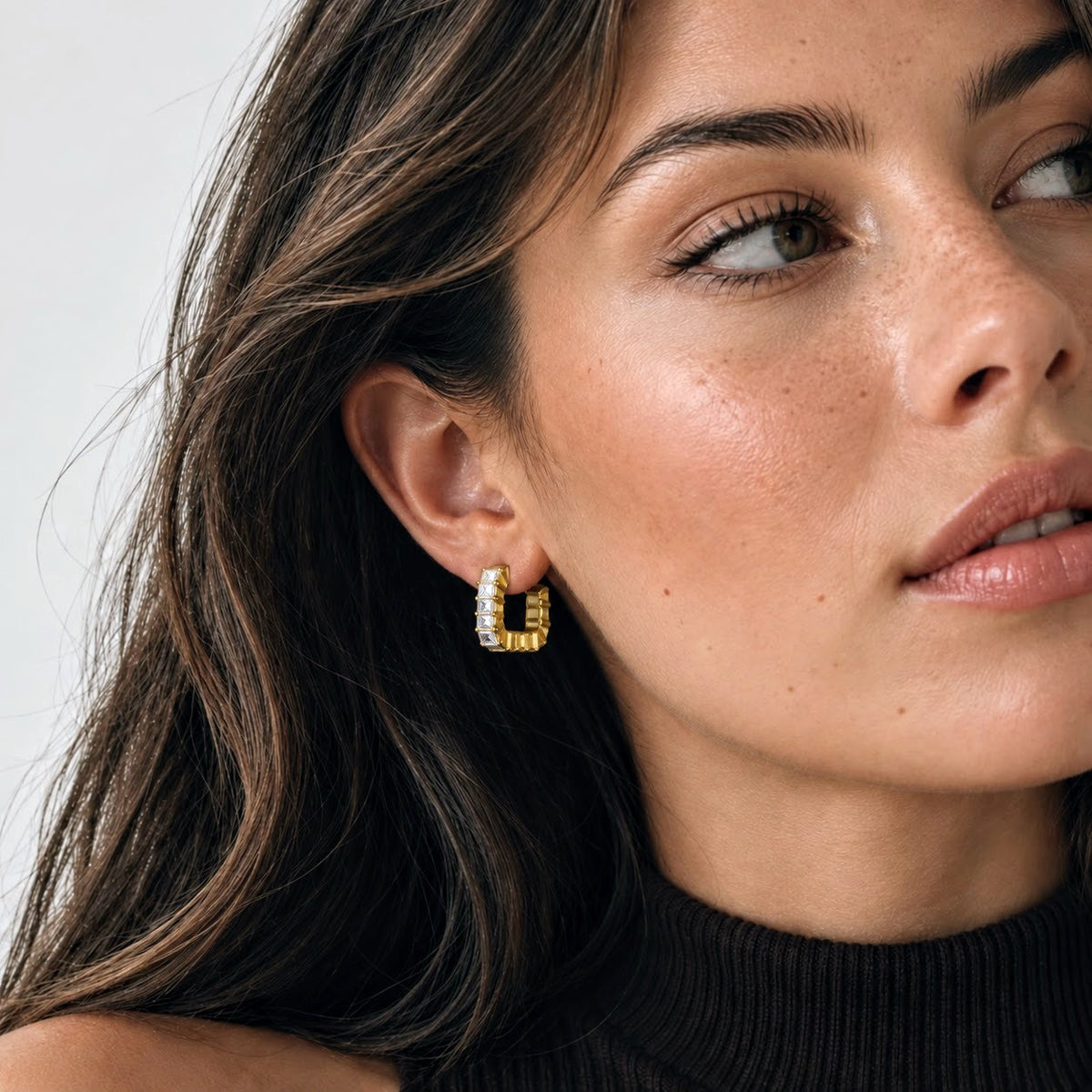 A close-up of a woman wearing a small gold huggie hoop earring with clear stones, shown against a light neutral background.