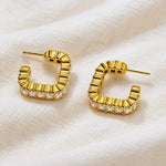 A pair of gold hoop-style stud earrings with a textured, segmented curved design and a row of clear stones along the front, displayed on a soft white textured fabric background.