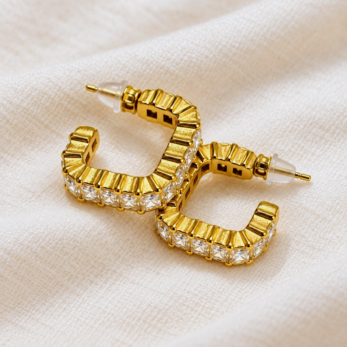 A pair of chunky gold hoop earrings embellished with clear square-cut crystals, resting on a textured off-white fabric background.