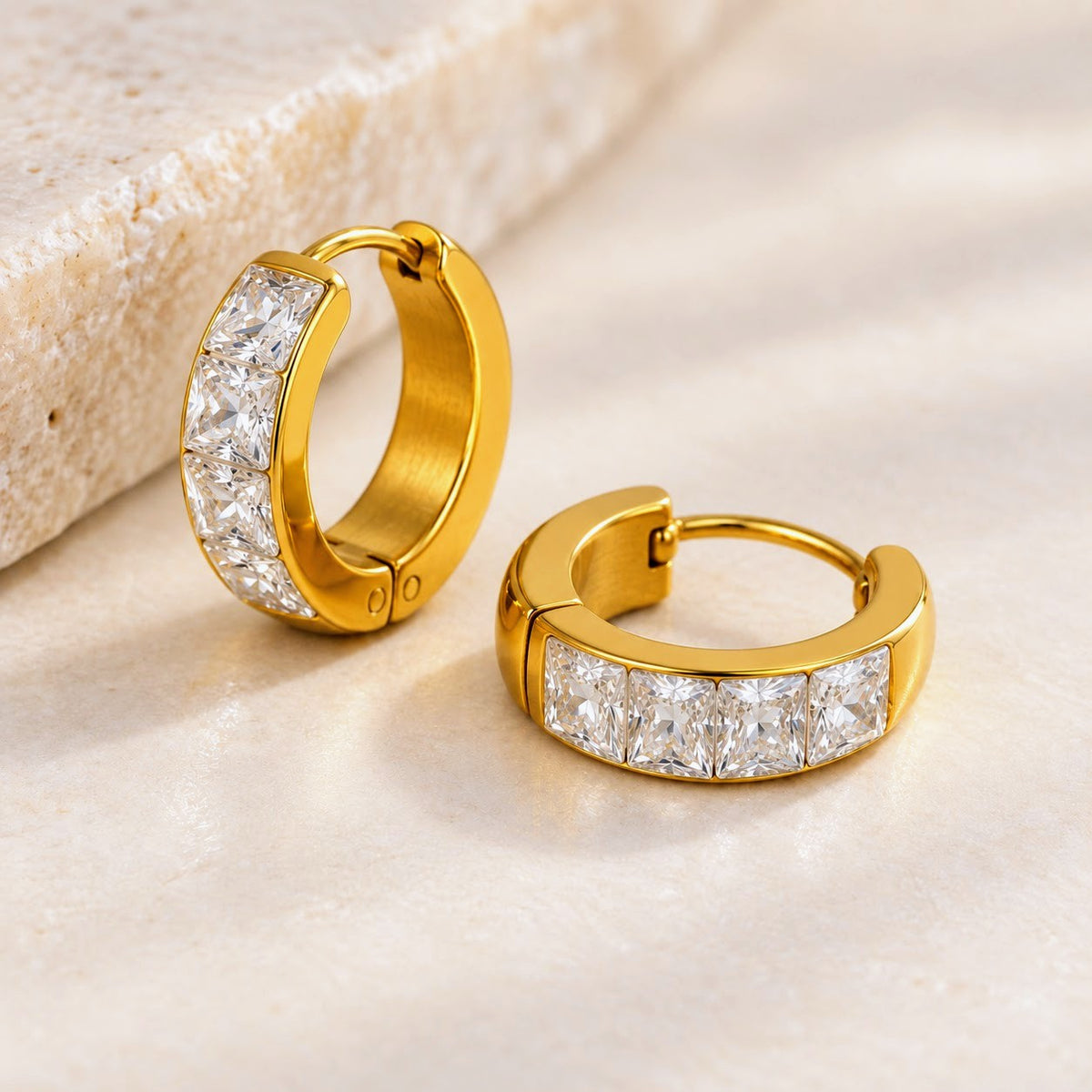 A pair of gold huggie hoop earrings with a channel-set row of clear square-cut stones, displayed on a light beige textured surface.