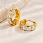 A pair of gold huggie hoop earrings with a channel-set row of clear square-cut stones, displayed on a light beige textured surface.