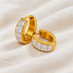 A pair of gold huggie hoop earrings paved with clear square-cut stones, resting on a soft, textured cream fabric background.