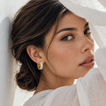 A close-up lifestyle shot of a woman wearing a small gold huggie hoop earring set with clear pavé stones, styled with a white sheer fabric backdrop.