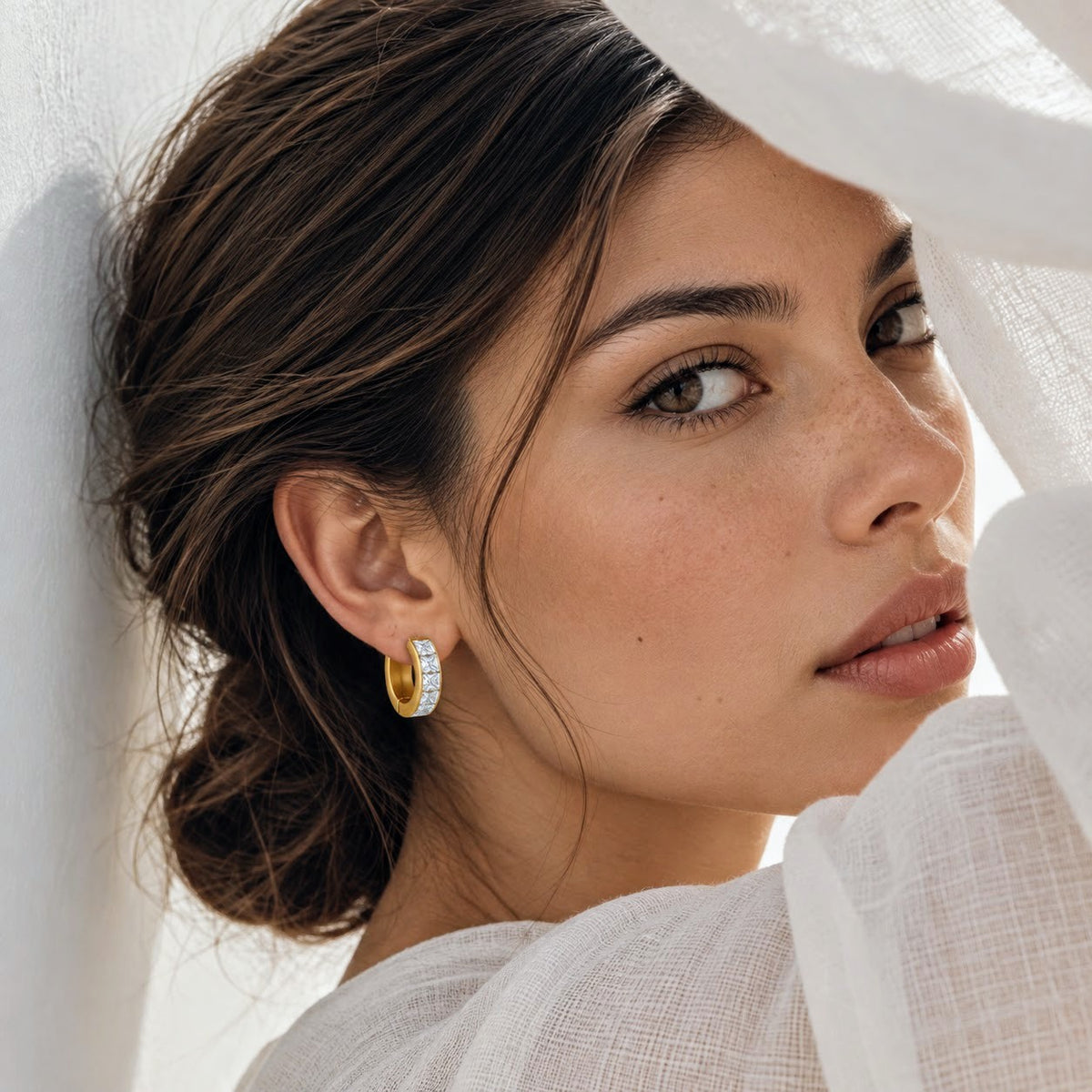 A close-up lifestyle shot of a woman wearing a small gold huggie hoop earring set with clear pavé stones, styled with a white sheer fabric backdrop.