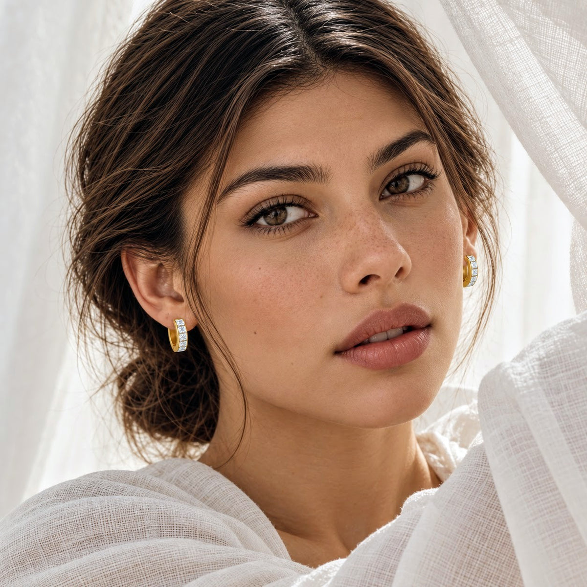 A close-up portrait of a woman wearing small gold huggie hoop earrings with clear gemstone accents, styled with a white textured top and sheer white drapery.