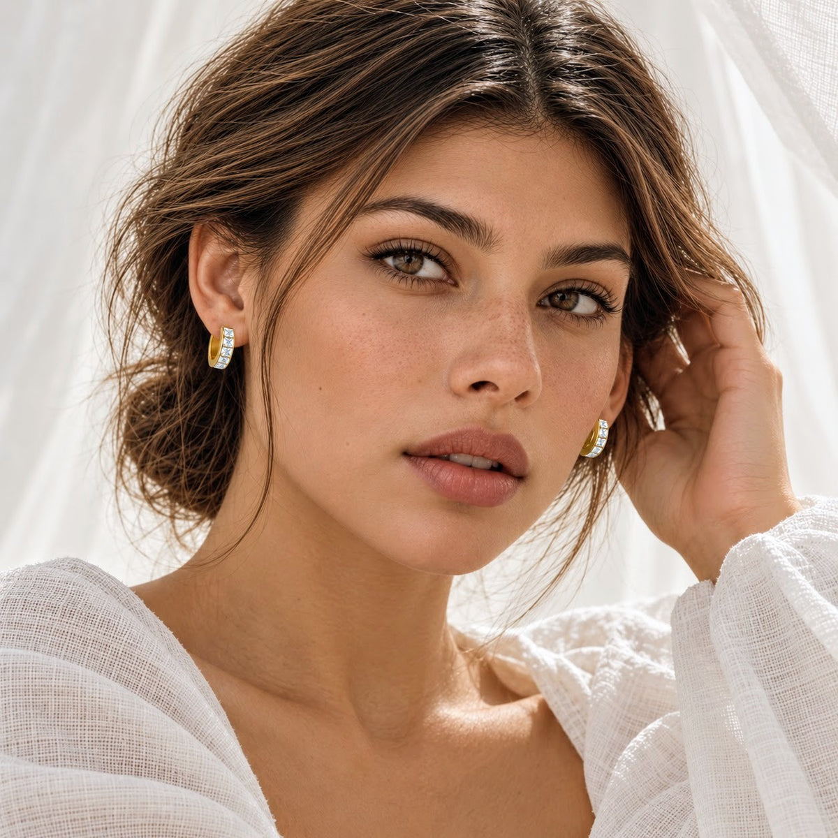 A close-up portrait of a woman wearing small gold huggie hoop earrings with clear gemstones, set against a soft white draped fabric background.