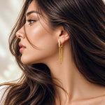 A close-up side profile of a woman with long dark brown hair wearing a delicate gold huggie hoop earring with dangling chain elements.