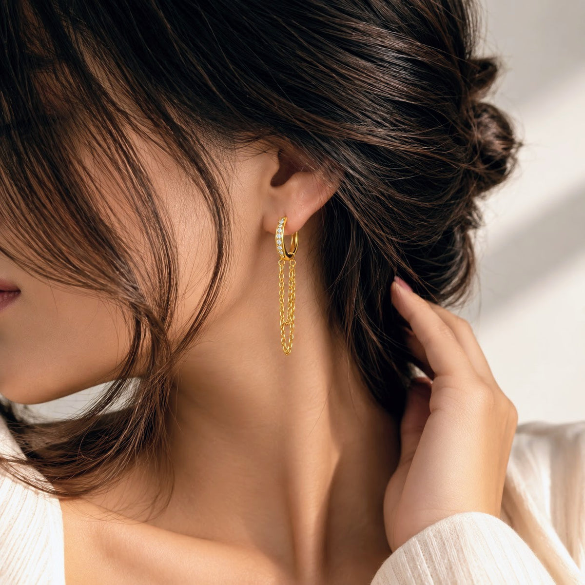 A close-up lifestyle shot of a woman wearing a delicate gold huggie hoop earring with dangling chain and pearl accents.