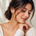 A close-up lifestyle shot of a woman wearing delicate gold dangle earrings, resting her chin on her hand against a soft, warm neutral background.