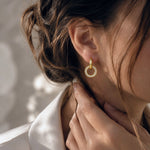 A close-up lifestyle shot of a woman wearing a delicate gold hoop earring adorned with small clear crystals, set against a softly lit, neutral background.