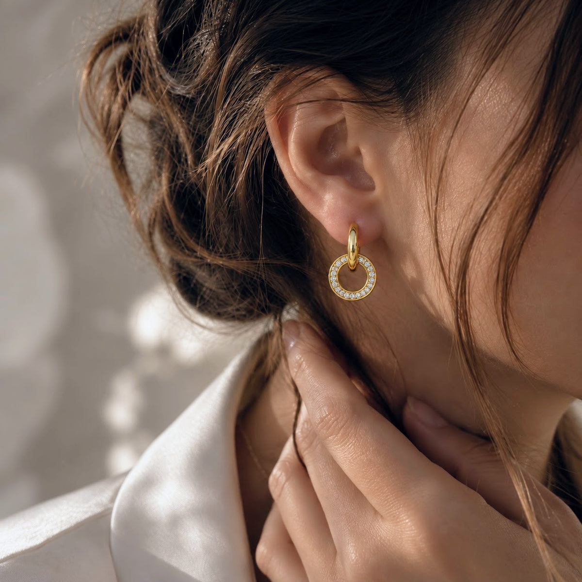 A close-up lifestyle shot of a woman wearing a delicate gold hoop earring adorned with small clear crystals, set against a softly lit, neutral background.