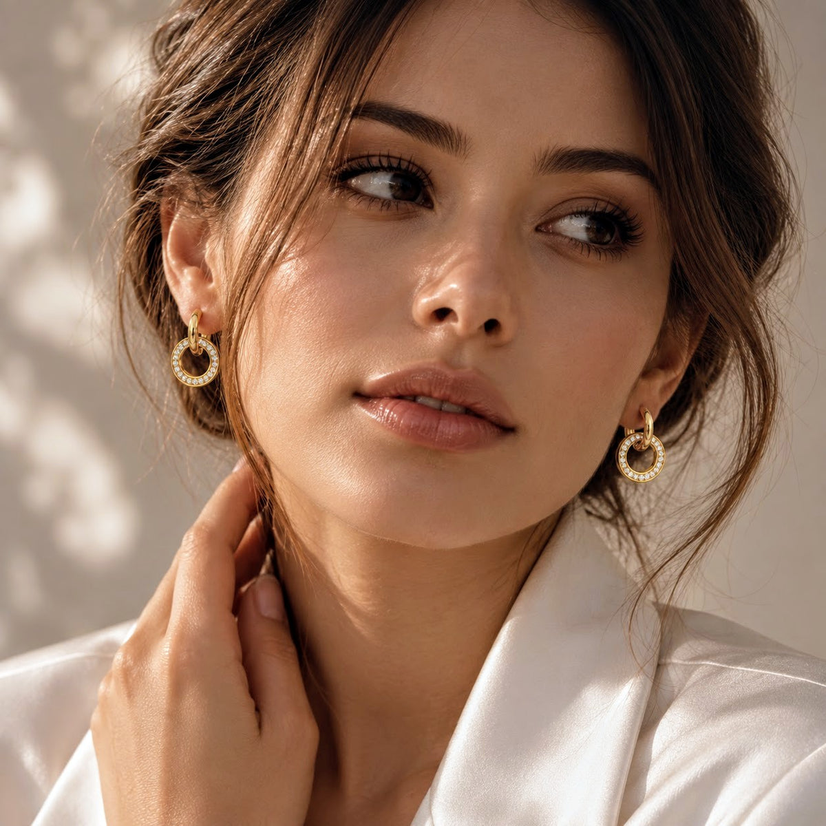 A close-up of a woman wearing small gold hoop earrings with a textured, beaded outer edge. She is looking off to the side with a soft, warm expression, wearing a white collared shirt against a softly lit beige background.