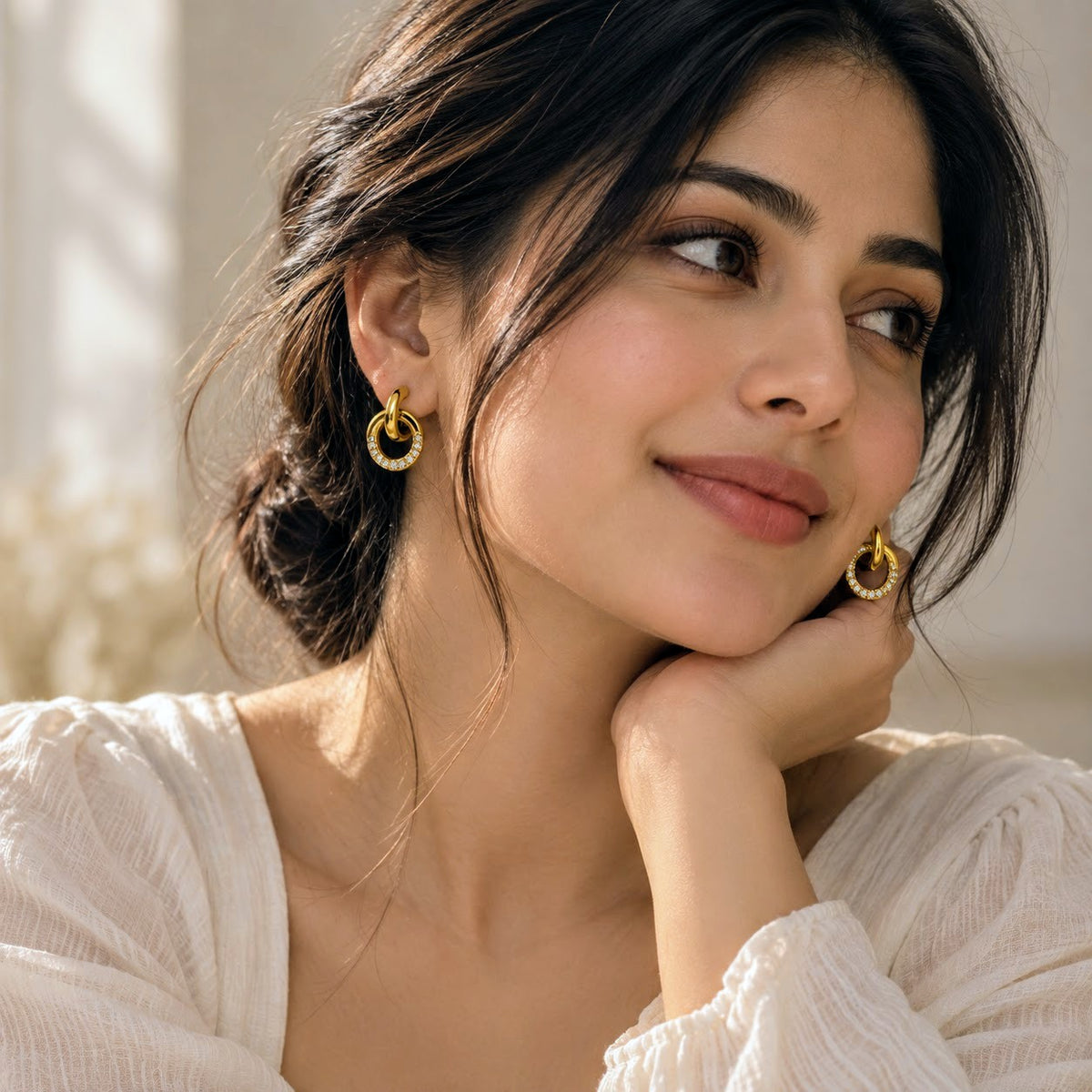 A woman with dark hair in a low bun wearing small gold hoop earrings with clear stones, posing in a white blouse against a softly lit neutral background.