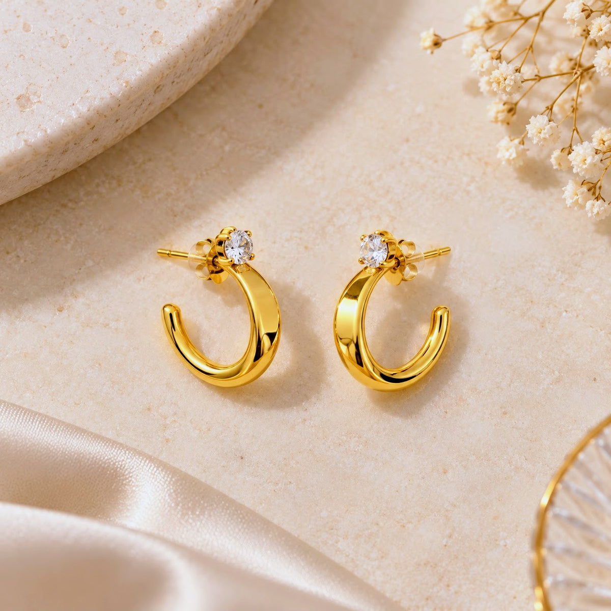 A pair of gold crescent-shaped hoop earrings with a small clear round gemstone in a floral setting, displayed on a textured  background.