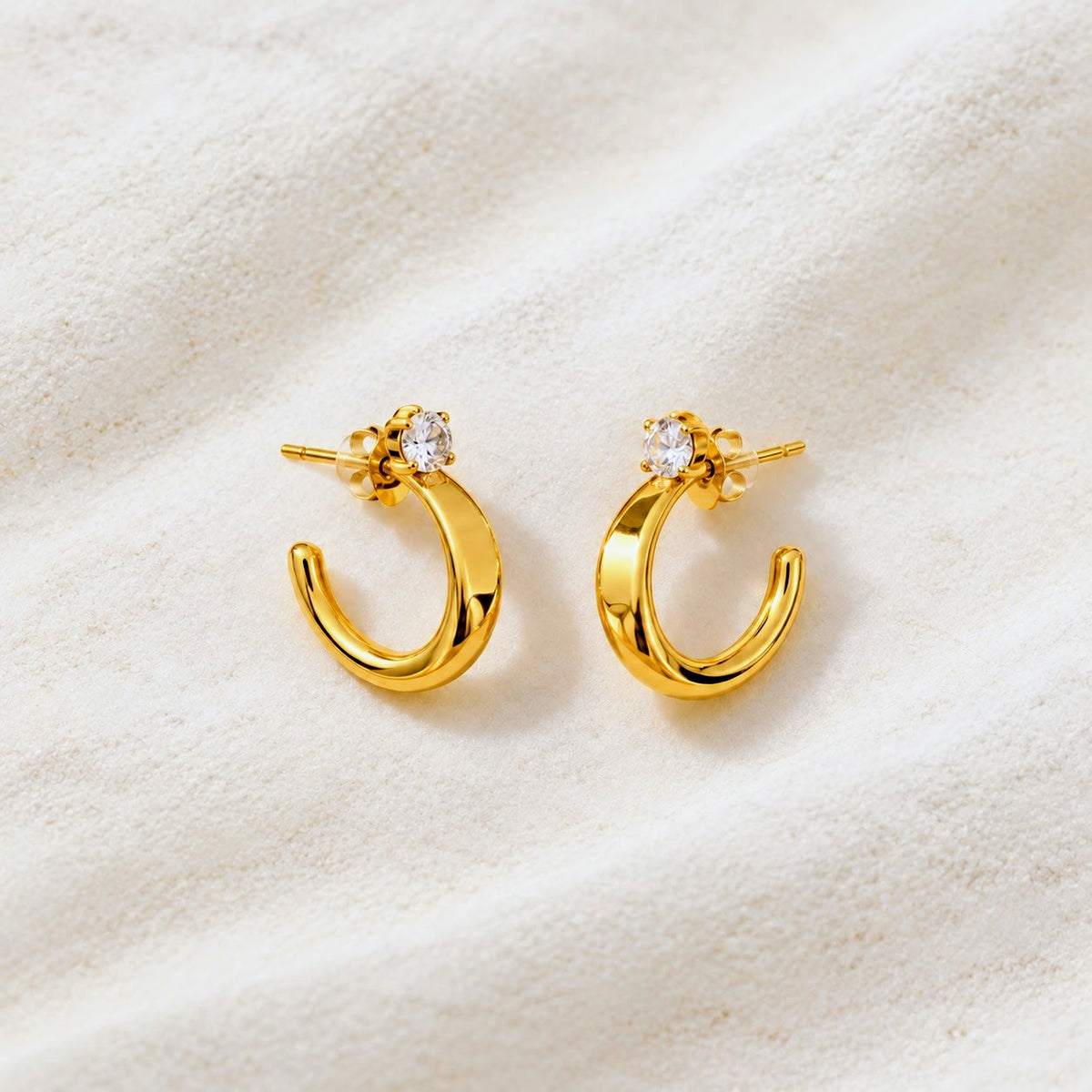 A pair of gold crescent-shaped hoop earrings with a small clear round gemstone in a floral setting, displayed on a textured cream fabric background.