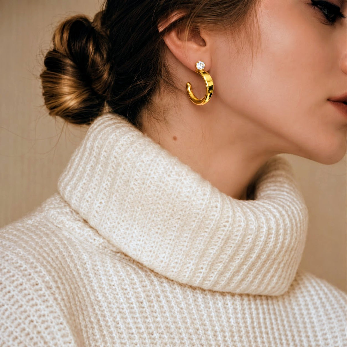 A close-up of a woman wearing gold crescent-shaped hoop earrings with small clear stones, paired with a cream-colored knit turtleneck sweater against a warm beige background.