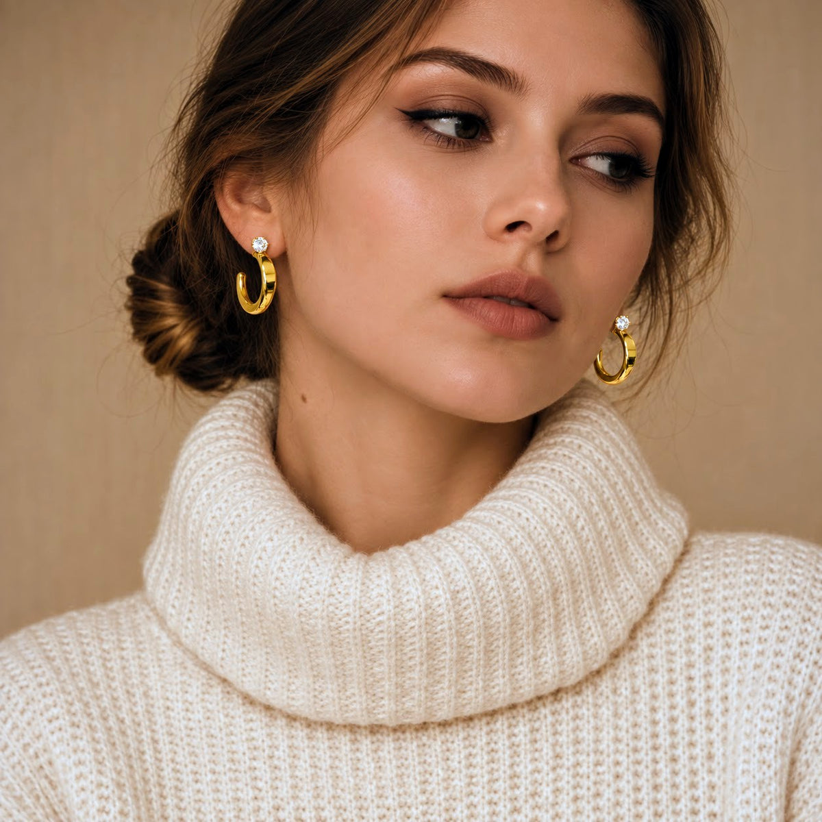 A close-up of a woman wearing gold crescent-shaped hoop earrings with small white pearl accents, paired with a cream-colored knit turtleneck sweater against a warm beige background.