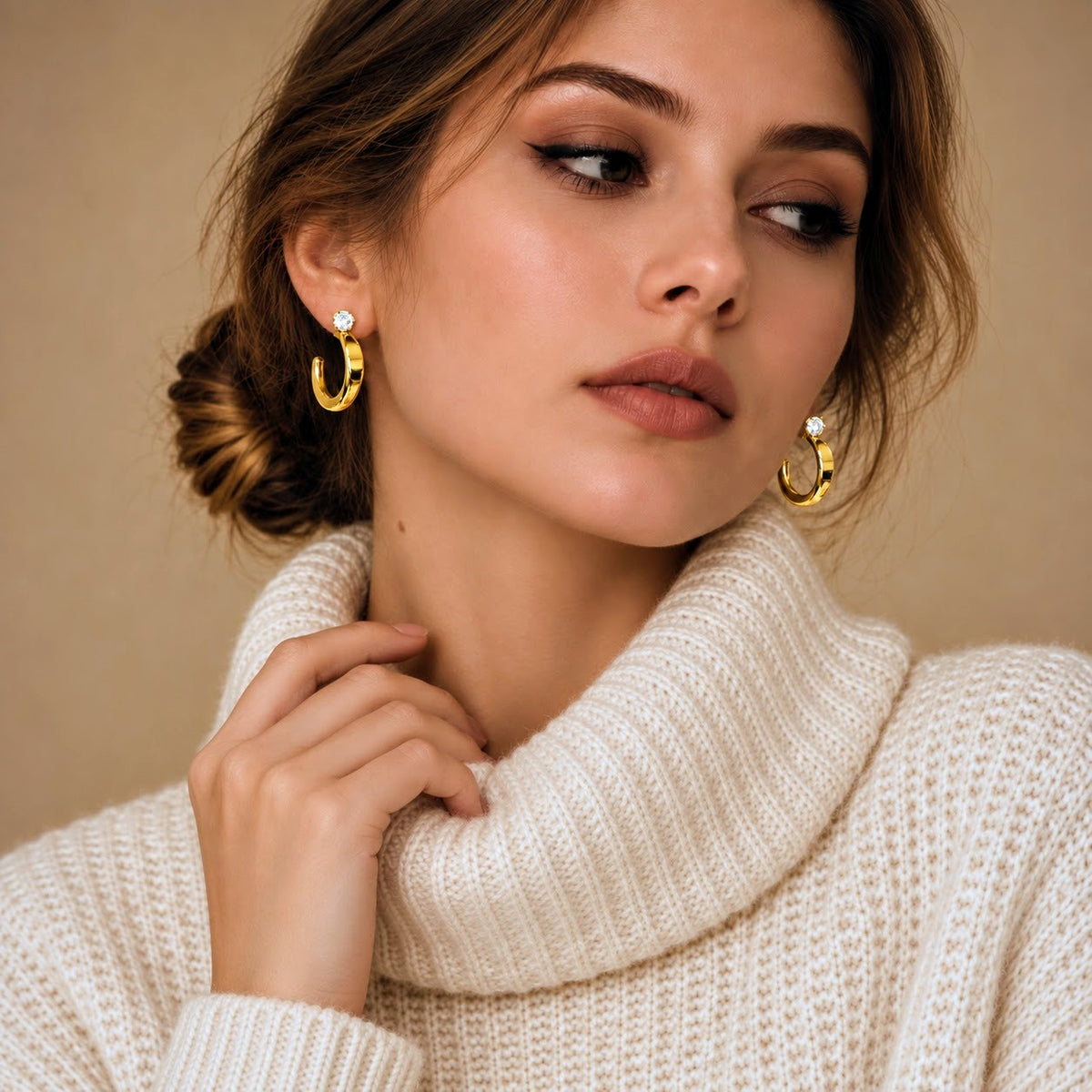 A close-up of a woman wearing gold crescent-shaped hoop earrings with small clear stones, paired with a cream-colored knit turtleneck sweater against a warm beige background.
